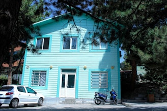 A turquoise-painted wooden house with two floors is surrounded by tall pine trees. The house has white-framed windows and a front door with a small set of stairs leading up to it. In front of the house, a white car and a blue scooter are parked on a paved area.