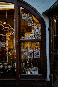 A shop window display featuring various small, intricate trinkets and ornaments. These decorations are colorful and include miniature scenes and figures possibly representing traditional or cultural themes. The window is framed by dark wood, and soft warm lighting creates a cozy and inviting atmosphere.