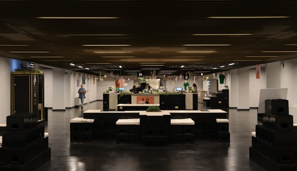 A modern interior space with a large central counter, adorned with plants and various decorations. On either side, there are seating arrangements made of stacked black blocks with white cushions. The ceiling has a wooden panel design and the floor is polished, adding a sleek look. Various shirts and banners hang on the walls, and there are a few people in the background near the counter.