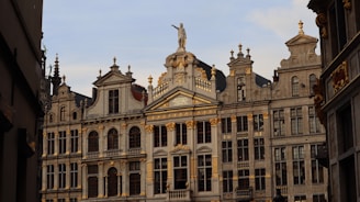 A historic building adorned with ornate architectural details, featuring multiple arched windows and decorative gold accents. The structure displays elements of classical architecture, including columns and a statue atop the roof.