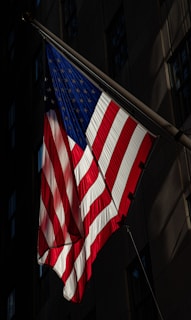 An American flag prominently displayed, with its stars and stripes clearly visible. It is set against a dark building façade in the background, with light casting a dramatic shadow.