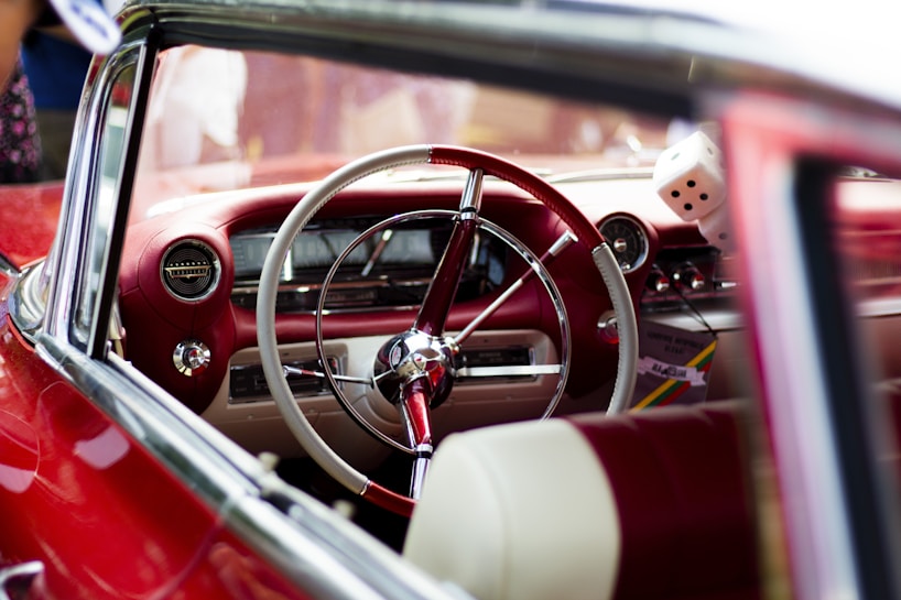 A vintage car interior featuring a red and white steering wheel, classic dashboard, and retro details. The dashboard includes an old-style radio, speedometer, and a pair of fuzzy dice hanging from the rear-view mirror.