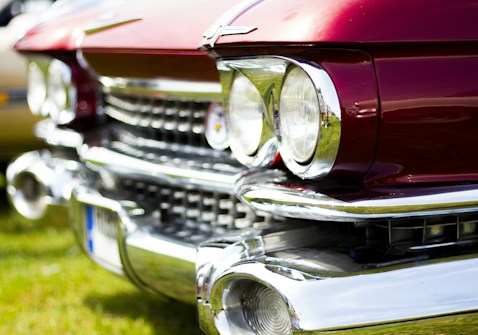A close-up of a classic car's front end, featuring prominent round headlights and a shiny chrome grille. The car's body is a deep red color with an intricate design that includes chrome accents. The background is slightly blurred with patches of green grass, indicating a possible outdoor setting.