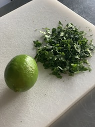 A lime and a pile of chopped green herbs are placed on a white cutting board. The background appears to be a stainless steel surface, suggesting a kitchen setting.