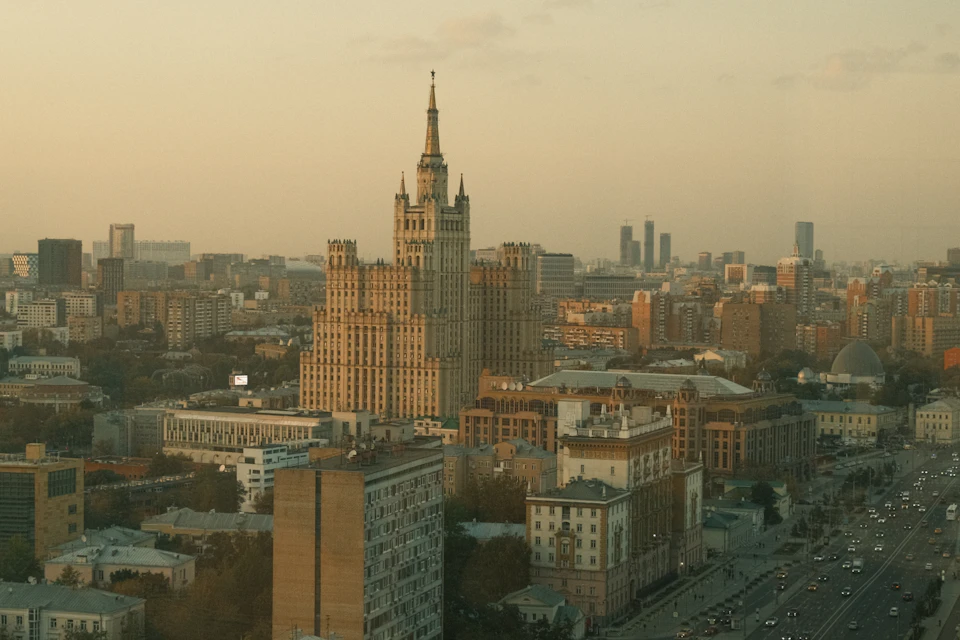 The image presents a panoramic view of the Moscow skyline, dominated by the majestic Kremlin Tower and the historic St. Basil's Cathedral. The tower stands tall against an orange sky, its spire piercing through the clouds. The cathedral, with its distinctive onion-like shape, is visible in the background. The cityscape is a blend of architectural styles, showcasing the rich history and cultural heritage of Moscow. The buildings are bathed in warm hues, reflecting off their surfaces under the soft light of dusk or dawn.