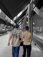 A couple is walking on a city sidewalk under a series of large awnings. One person is wearing a Hawaiian-style shirt and a blue cap, while the other is in a tan shirt. The background is monochrome, emphasizing the colorful details of the shirts and cap.