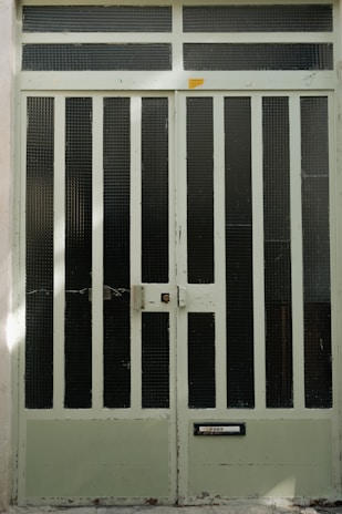 A large metal door painted in a pale green color, featuring vertical segments with mesh screens. The door has a weathered look, with scratches and paint peeling in some areas. A small rectangular mail slot labeled 'POST' is located near the bottom, and a doorbell or intercom is affixed near the center.