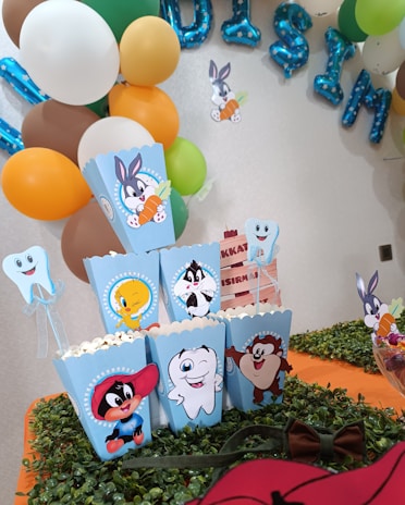 A festive party setup featuring colorful balloons in the background. Brightly colored popcorn boxes showcase cartoon characters, including a rabbit with a carrot and other well-known animated figures. The table is adorned with a green leafy runner and additional decorations such as a tooth-shaped figure and a bowl of candy.