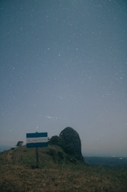 A nighttime landscape with a prominent rock formation in the background, illuminated by a clear, star-filled sky. In the foreground, a sign reads 'El Salvador' amidst grassy terrain.