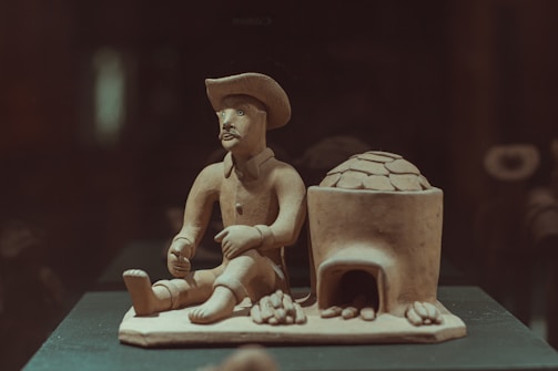 A clay sculpture depicts a seated figure wearing a hat, holding an object in one hand. Next to the figure is a clay oven or structure with an arched opening and several smaller objects at its base, possibly resembling bread or pastries.