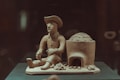 A clay sculpture depicts a seated figure wearing a hat, holding an object in one hand. Next to the figure is a clay oven or structure with an arched opening and several smaller objects at its base, possibly resembling bread or pastries.