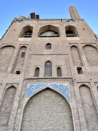 A large, ancient brick structure with tall arches and intricate tile work featuring blue patterns. The facade is detailed, showcasing historical architecture with several windows and an arched entry that is partially blocked. The building seems to be undergoing restoration work.