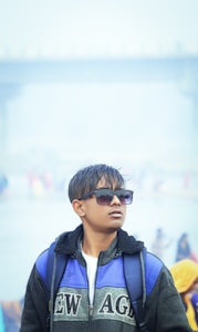A young person wearing sunglasses stands confidently outdoors. They have short, wet hair and are dressed in a jacket with the text 'NEW AGE' visible. A backpack is strapped on their shoulders. The background appears to be misty or foggy, with blurred figures and a structure visible.