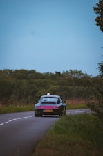Estate car driving along a tree-lined European countryside road under a clear sky.