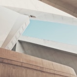 A modern architectural structure with angular lines and large concrete surfaces. The composition features intersecting planes and a clear blue sky visible through open spaces. The design is minimalistic, with smooth textures and subtle shading.