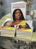 A stack of books titled 'In Love with the Possibilities' with a close-up of one prominently displayed on top. The cover features a photograph of a woman smiling, with additional copies visible underneath. There is glassware and a spool of twine in the background on a reflective tabletop.
