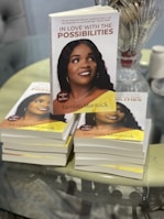 A stack of books titled 'In Love with the Possibilities' with a close-up of one prominently displayed on top. The cover features a photograph of a woman smiling, with additional copies visible underneath. There is glassware and a spool of twine in the background on a reflective tabletop.