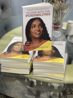 A stack of books titled 'In Love with the Possibilities' with a close-up of one prominently displayed on top. The cover features a photograph of a woman smiling, with additional copies visible underneath. There is glassware and a spool of twine in the background on a reflective tabletop.
