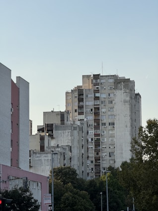 Multiple high-rise residential buildings with a worn and aged appearance are shown. The structures are mostly gray and white, surrounded by green trees under a clear blue sky.
