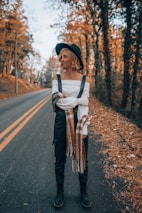 A person stands on a quiet, leaf-strewn road lined with trees. They are dressed in a stylish off-the-shoulder white top, dark overalls, a scarf, boots, and a black hat. The leaves on the ground and trees indicate an autumn setting.