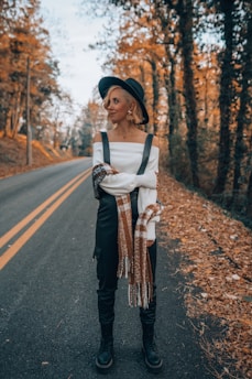A person stands on a quiet, leaf-strewn road lined with trees. They are dressed in a stylish off-the-shoulder white top, dark overalls, a scarf, boots, and a black hat. The leaves on the ground and trees indicate an autumn setting.
