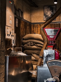 Inside a dimly lit room with a rustic and industrial feel, sacks are stacked against a wooden panel wall. A red industrial machine with a large pipe protrudes into the space, alongside a metallic control panel on the wall with warning signs. Nearby, a weighing scale and other equipment suggest a functional, possibly coffee-related, workspace. A portrait of a man hangs on the upper wall, hinting at a historical or personal significance.