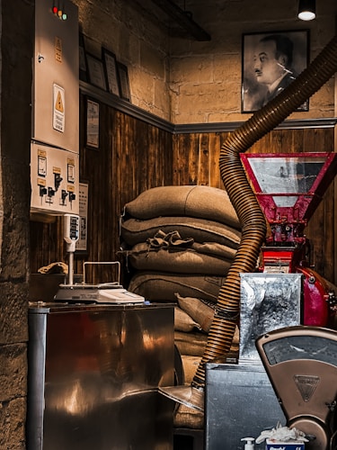 Inside a dimly lit room with a rustic and industrial feel, sacks are stacked against a wooden panel wall. A red industrial machine with a large pipe protrudes into the space, alongside a metallic control panel on the wall with warning signs. Nearby, a weighing scale and other equipment suggest a functional, possibly coffee-related, workspace. A portrait of a man hangs on the upper wall, hinting at a historical or personal significance.