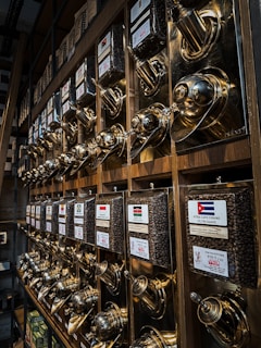 A wall display filled with shiny, metallic coffee dispensers. Each dispenser is labeled with different country flags and names, filled with coffee beans. The dispensers have a polished, brass-like finish and are organized in a neat, grid-like fashion. The setting suggests a specialty coffee shop or a store focused on various international coffee blends.