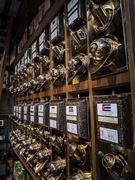 A wall display filled with shiny, metallic coffee dispensers. Each dispenser is labeled with different country flags and names, filled with coffee beans. The dispensers have a polished, brass-like finish and are organized in a neat, grid-like fashion. The setting suggests a specialty coffee shop or a store focused on various international coffee blends.