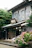 A traditional Japanese building with a wooden exterior and a tiled roof is surrounded by lush greenery. In front of the building, there is a vibrant display of blooming pink and purple hydrangeas. The architecture is characterized by sliding wooden doors and a rustic sign, indicating an old-fashioned and peaceful setting.