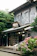 A traditional Japanese building with a wooden exterior and a tiled roof is surrounded by lush greenery. In front of the building, there is a vibrant display of blooming pink and purple hydrangeas. The architecture is characterized by sliding wooden doors and a rustic sign, indicating an old-fashioned and peaceful setting.
