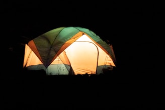 A warmly lit tent with a combination of green, orange, and beige fabric set against a dark background, likely indicating nighttime. The inside of the tent glows, providing a cozy and inviting atmosphere.
