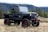 A rugged, black off-road vehicle with large tires and red accents parked on grassy terrain. It is an open-top design, with no doors or roof, and features a winch on the front bumper. In the background, there are trees and a mountain range under a cloudy sky.
