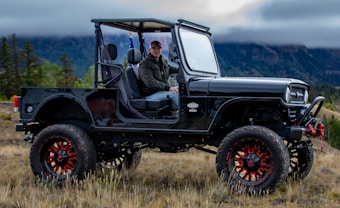 A person is sitting in a lifted black off-road vehicle with large red and black wheels, parked in a grassy area with cloudy mountain scenery in the background. The vehicle has an open frame, and the person appears to be wearing a cap and jacket.