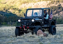 A rugged off-road vehicle with a black exterior is parked in a natural setting surrounded by grass and trees. There are two people in the vehicle, and an American flag is mounted on the vehicle's rear. The background shows a forested area with hills, suggesting a scenic outdoor location.