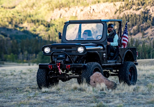 A rugged off-road vehicle with a black exterior is parked in a natural setting surrounded by grass and trees. There are two people in the vehicle, and an American flag is mounted on the vehicle's rear. The background shows a forested area with hills, suggesting a scenic outdoor location.