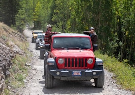 A group of off-road vehicles, led by a red jeep with a license plate reading 'TOWN & COUNTRY', is driving down a dirt path surrounded by lush greenery. Two people are seen interacting on the red jeep, one wearing a plaid shirt and a hat, while the other is mostly obscured by their hand. The dirt road appears rugged, indicating an adventurous journey or off-road trail.