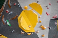 An indoor climbing wall featuring an arrangement of colorful holds and geometric shapes. Dominant yellow panels with various smaller holds in colors such as red, blue, green, and brown are scattered across the textured surface.