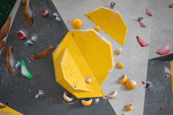 An indoor climbing wall featuring an arrangement of colorful holds and geometric shapes. Dominant yellow panels with various smaller holds in colors such as red, blue, green, and brown are scattered across the textured surface.