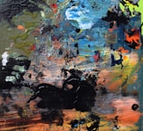 An abstract painting featuring a chaotic blend of textures and colors. Brush strokes and splatters in shades of blue, green, orange, and black create a dynamic and energetic composition. The surface is filled with layers of paint, creating a sense of depth and complexity.