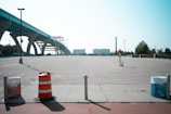 A large, empty parking lot stretches into the distance with a few scattered light poles and minimal vegetation. In the foreground, there are an orange traffic barrel and two waste bins, one with a red cover and one with a blue cover. To the left, an elevated highway with structural arches extends across the scene. In the background, an industrial building with a tall chimney and some trees are visible.