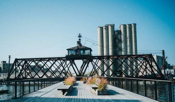 An industrial scene featuring a rusty iron bridge with a central control house, set against large cylindrical silos. The foreground includes a boardwalk with planters containing tall grasses. The sky is clear and blue, contributing to an open and expansive atmosphere.