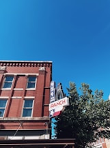 A red brick building with several windows and a prominent retro-style neon sign featuring the text '95.9 FM RANCH'. The upper section of the building is highlighted, with a bright blue sky in the background. A partial green tree is visible on the right.