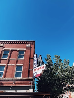 A red brick building with several windows and a prominent retro-style neon sign featuring the text '95.9 FM RANCH'. The upper section of the building is highlighted, with a bright blue sky in the background. A partial green tree is visible on the right.