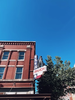 A red brick building with several windows and a prominent retro-style neon sign featuring the text '95.9 FM RANCH'. The upper section of the building is highlighted, with a bright blue sky in the background. A partial green tree is visible on the right.