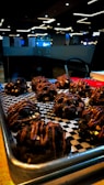 A tray of chocolate cookies drizzled with caramel glaze is placed on a checkered black and white paper. The cookies are chunky, with visible chocolate chips and a shiny caramel topping. The setting appears to be a modern cafe with dim lighting and contemporary decor, seen in the background.