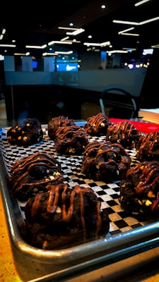 A tray of chocolate cookies drizzled with caramel glaze is placed on a checkered black and white paper. The cookies are chunky, with visible chocolate chips and a shiny caramel topping. The setting appears to be a modern cafe with dim lighting and contemporary decor, seen in the background.