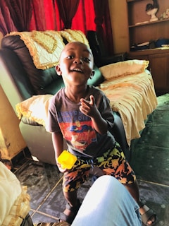 A young child is standing and smiling in a cozy living room. The child is wearing a colorful t-shirt with cartoon graphics and patterned shorts. There is a yellow toy or cup in one of the child's hands. The room has a leather couch with decorative pillows and a blanket. Red curtains are drawn open behind the couch. There is a bookshelf with various items on it in the background.