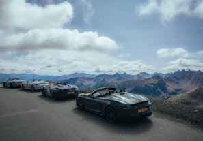 A panoramic view of the Dolomites with a lineup of exotic supercars parked on a scenic overlook.