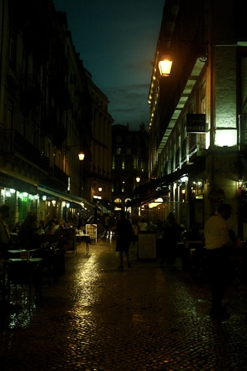 A cobblestone street at night lined with buildings, dimly lit by street lamps. Sidewalk cafes with tables visible, some occupied by people. The atmosphere is calm and laid-back, with a hint of liveliness from the activity in the cafes.